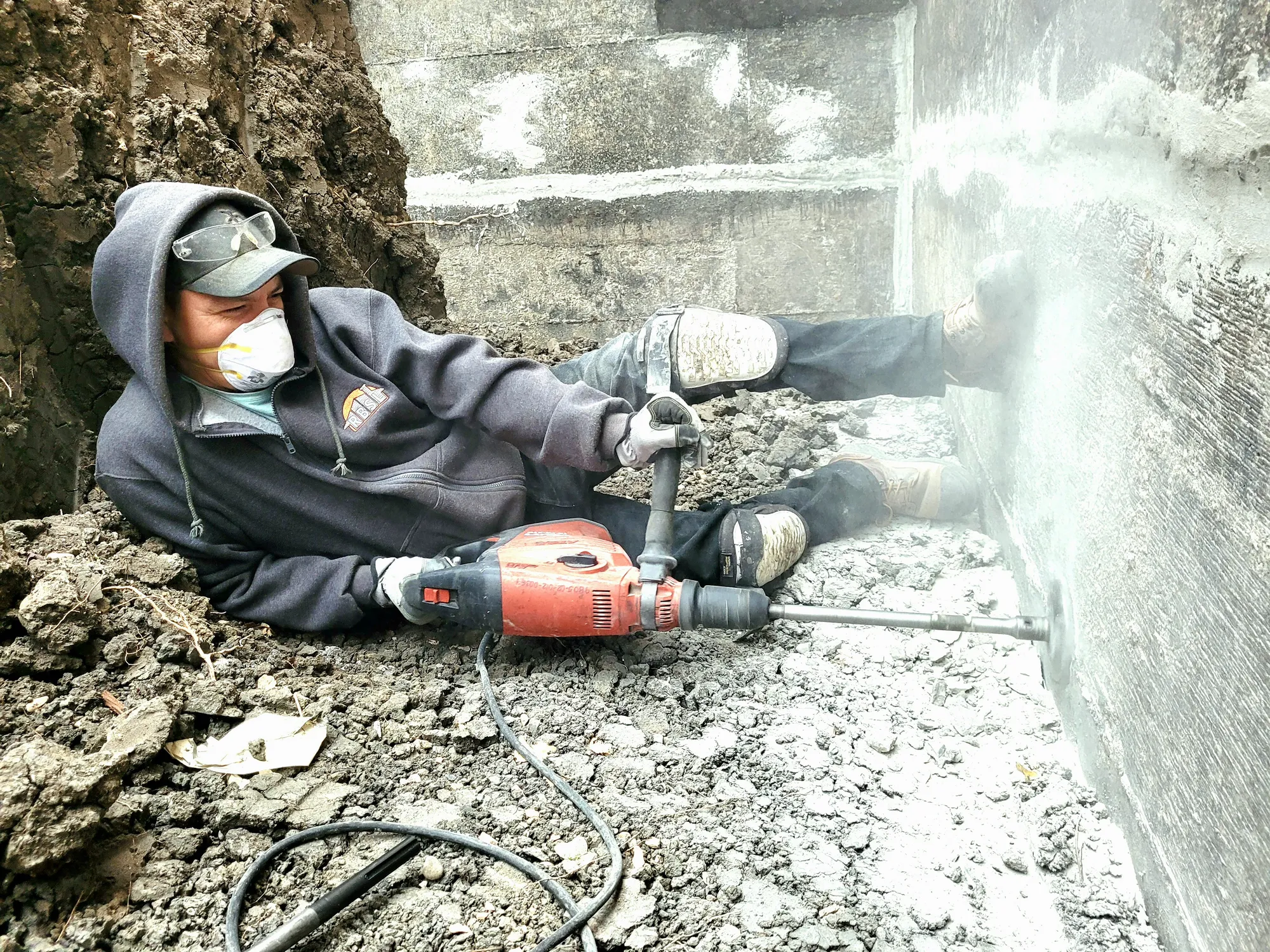 A man laying in a trench drilling a hole into a concrete wall